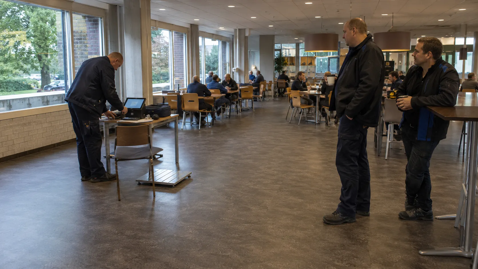 Een eenvoudige opstelling in de hoek van een bedrijfskantine: een tafel met apparatuur, een meetplaat op de vloer en enkele wachtende collega\'s.